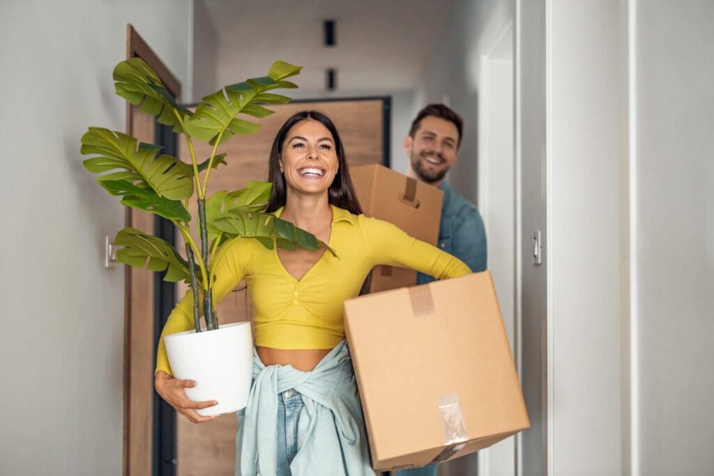 A joyful couple moving into their new home at Windsor Place in Beavercreek, Ohio, with the woman holding a plant and the man carrying a cardboard box.